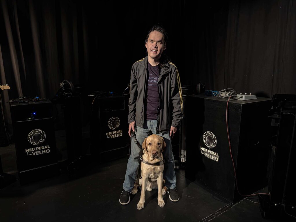 Marcelo posando para foto no centro do teatro do evento com Quim, seu cão guia, também posando e posicionado entre suas pernas.