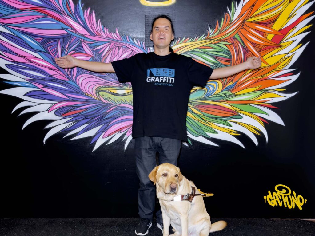 Marcelo de braços abertos em frente do graffiti acessível que representa asas estilizadas com uma auréola sobre a cabeça e sei companheiro cão-guia, Quim, em sua frente.